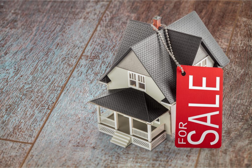 Miniature house with a red "For Sale" tag on a wooden floor.