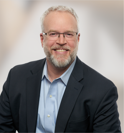 Man with gray hair and beard wearing glasses and dark suit smiling
