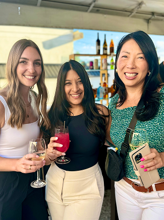 Three women smiling at an outdoor event while holding colorful drinks.