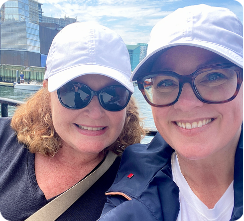 Two women in white caps smiling while sitting outdoors near the water.