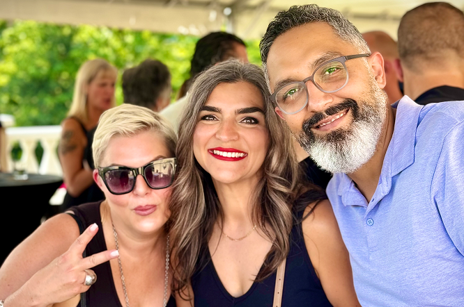 Three friends smiling outdoors at a daytime gathering, with one showing a peace sign.