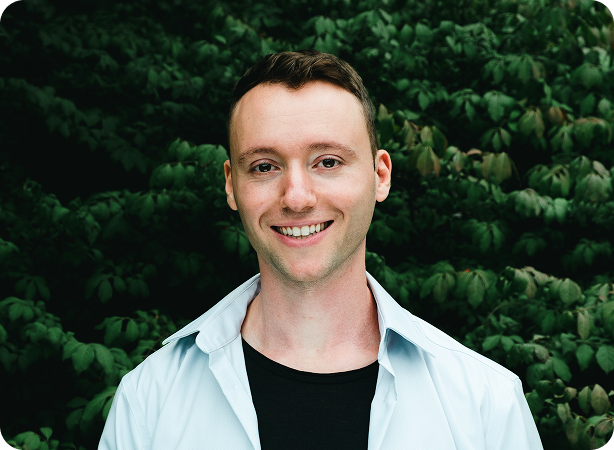 Smiling man standing outdoors in front of dense green foliage.