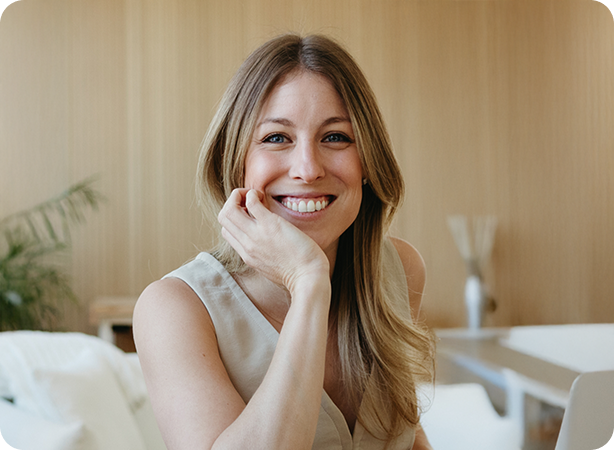 Smiling woman sitting indoors with her hand resting on her chin.